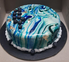 White round celebration cake with smooth fondant frosting decorated with purple orchids, small white filler flowers, and scattered sugar pearls; placed on a gold cake board against a dark background. No visible text. Tone elegant and celebratory.